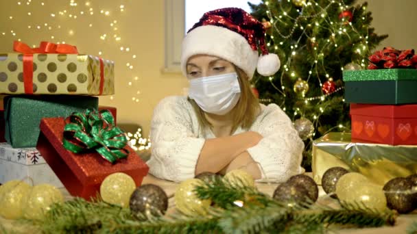Sorglig kvinna i en tomte hatt och en skyddande mask sitter ensam mot bakgrund av en julgran och lådor med presenter. Jul under COVID-19-pandemin. — Stockvideo