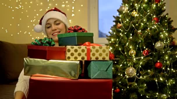 Mujer alegre en un sombrero de Santa entre los regalos preparados para los seres queridos. En el contexto de los comedores de Navidad. — Vídeo de stock