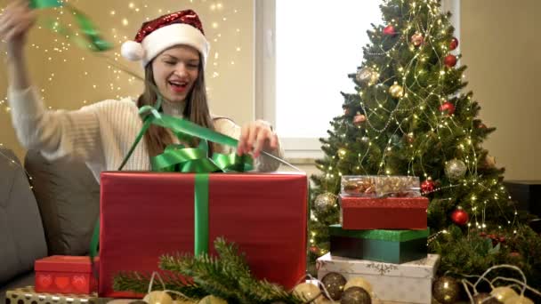 Bella donna bruna in un cappello di Babbo Natale è felicemente disimballando il suo regalo di Natale. Buon Natale. Ciao.. — Video Stock