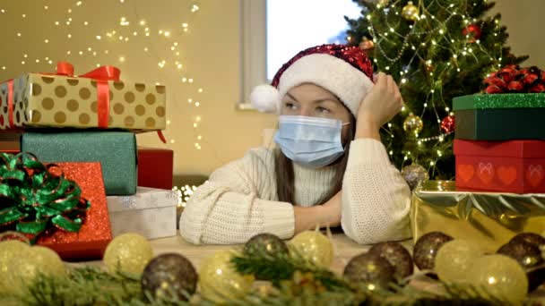 サンタの帽子の悲しい女性と保護マスクはクリスマスツリーとギフト付きの箱の背景に一人で座っています。COVID-19大流行中のクリスマス. — ストック動画