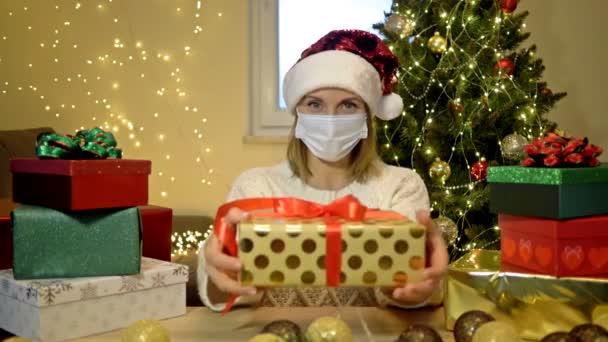 Mujer atractiva con sombrero de Santa y mascarilla muestra maravillosamente envuelto regalo. Sobre el fondo de un árbol de Navidad y cajas brillantes con regalos. Navidad a pesar de la pandemia de COVID-19. — Vídeo de stock