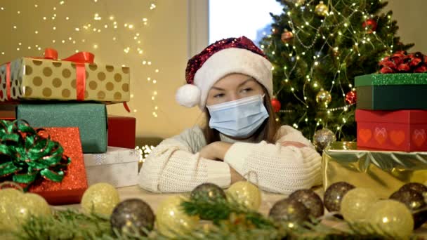 Mujer triste en un sombrero de Santa y una máscara protectora se sienta sola sobre el fondo de un árbol de Navidad y cajas con regalos. Navidad durante la pandemia de COVID-19. — Vídeo de stock