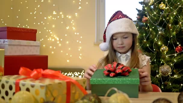 Niña de 6-7 años con un sombrero de Santa Claus sobre el fondo de un árbol de Navidad y cajas brillantes con regalos. Niño está muy decepcionado con el regalo de Navidad. — Vídeo de stock