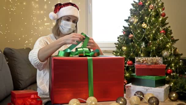 Hermosa mujer con un sombrero de Santa Claus y una máscara protectora en su cara está empacando regalos. En el fondo del árbol de Navidad. Navidad a pesar de la pandemia de COVID-19. — Vídeos de Stock