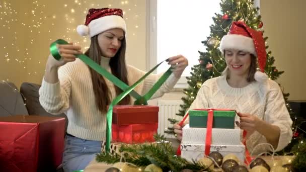 Dos adorables mujeres con sombreros de Santa están preparando regalos de Navidad. Sobre el fondo de un elegante árbol de Navidad. Humor festivo. — Vídeos de Stock