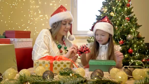 Mamá y su hija pequeña en los sombreros de Santa están preparando amorosamente regalos para sus seres queridos en el fondo de un árbol de Navidad. Humor festivo. Feliz Navidad.. — Vídeos de Stock