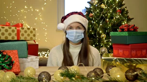 Trieste vrouw met een kerstmuts en een beschermend masker zit alleen tegen de achtergrond van een kerstboom en dozen met geschenken. Kerstmis tijdens de COVID-19 pandemie. — Stockvideo