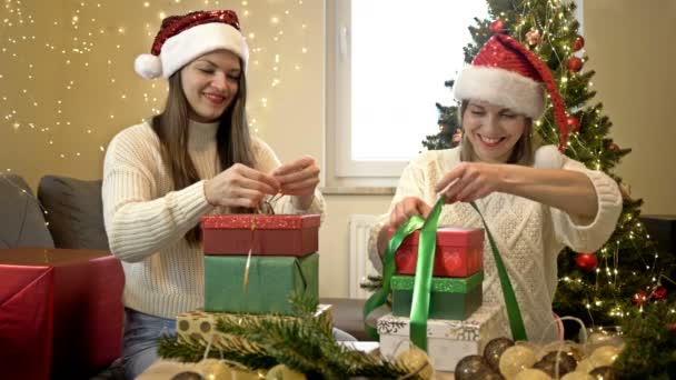Dos adorables mujeres con sombreros de Santa están preparando regalos de Navidad. Sobre el fondo de un elegante árbol de Navidad. Humor festivo. — Vídeos de Stock