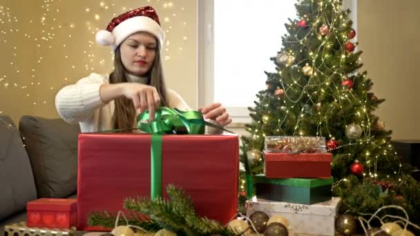 Hübsche brünette Frau mit Weihnachtsmütze packt fröhlich ihr Weihnachtsgeschenk aus. Frohe Weihnachten. — Stockvideo