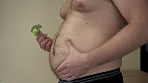 Dicke Mann zeigt die Falten des überschüssigen Fetts auf seinem Bauch mit einer Hand und hält Brokkoli mit der anderen. Problem mit Übergewicht und gesunder Ernährung. — Stockvideo