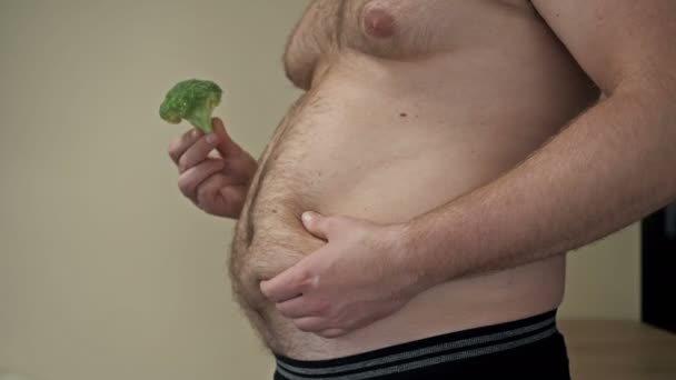脂肪の男は片手で彼の胃の上に余分な脂肪の折り目を示し、他のブロッコリーを保持しています。太りすぎで健康的な食事の問題. — ストック動画