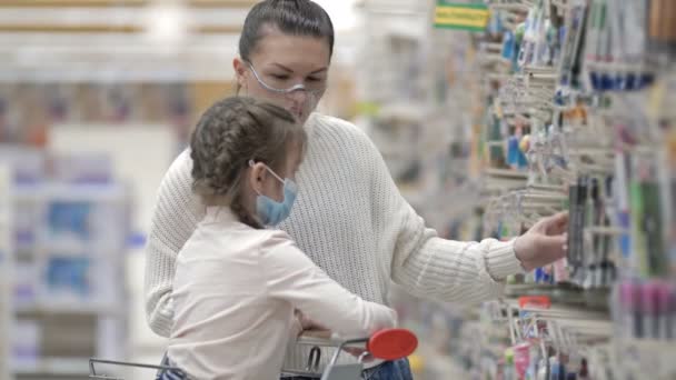 Koruyucu maskeler takan anne ve çocuk süpermarketten kırtasiye malzemesi seçiyorlar. Virüs salgını sırasında çocuklarla alışveriş yapmak. — Stok video