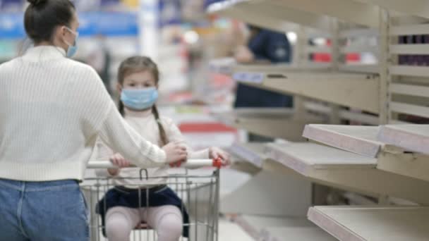 Een vrouw met een beschermend masker rolt een winkelwagentje langs de lege planken in de winkel. In een winkelwagen, een klein meisje met een beschermend masker. Handelsverstoringen als gevolg van de Covid-19-pandemie. — Stockvideo