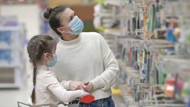 Koruyucu maskeler takan anne ve çocuk süpermarketten kırtasiye malzemesi seçiyorlar. Virüs salgını sırasında çocuklarla alışveriş yapmak. — Stok video