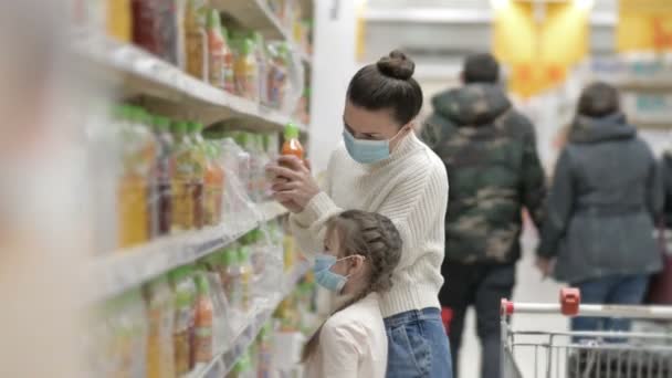 Mamá y su hija, de 6 a 7 años, eligen jugos en el supermercado. Ambos llevan máscaras protectoras. Compras con niños durante el brote del virus. — Vídeos de Stock