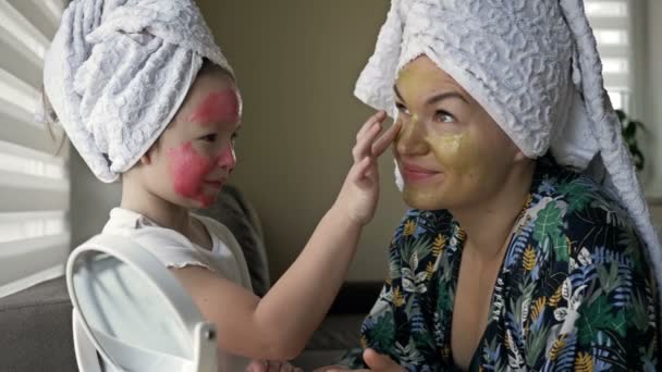 Une petite fille avec un masque cosmétique sur le visage aide sa mère à fabriquer un masque cosmétique. Heureux ensemble. — Video