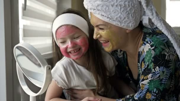 Молодая женщина с дочерью в возрасте 6-7 лет нанесла на их лица косметические маски. Они смотрят в зеркало и весело смеются.. — стоковое видео