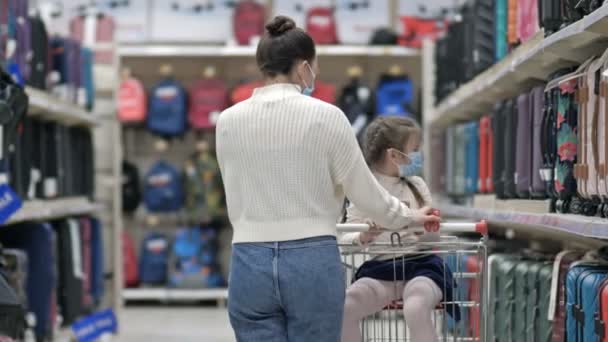 La mujer con su hija pequeña usando máscaras protectoras está eligiendo una maleta en un supermercado. Compras con niños durante el brote del virus. — Vídeos de Stock
