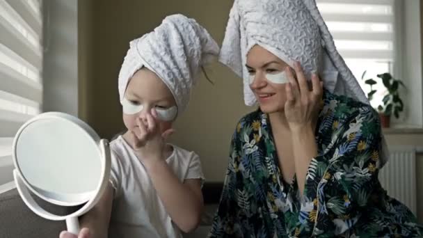 Schöne Mutter im Bademantel und ihre kleine Tochter zusammen mit Handtüchern auf dem Kopf und Maske auf dem Gesicht, während sie Wellness-Behandlungen genießt. — Stockvideo