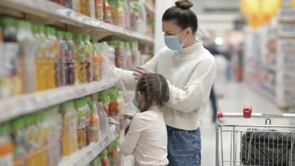Anya és lánya, 6-7 éves, válasszanak gyümölcslevet a szupermarketben. Mindkettő védőmaszkot visel. Vásárlás gyermekekkel a vírus kitörése alatt. — Stock videók