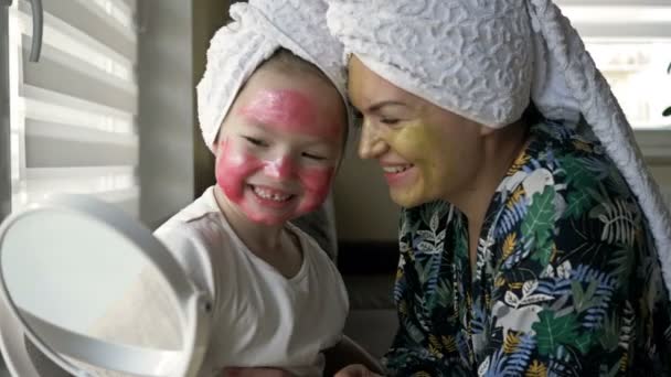 Jeune femme avec sa fille 6-7 ans appliqué des masques cosmétiques sur leurs visages. Ils se regardent dans le miroir et rient joyeusement. — Video