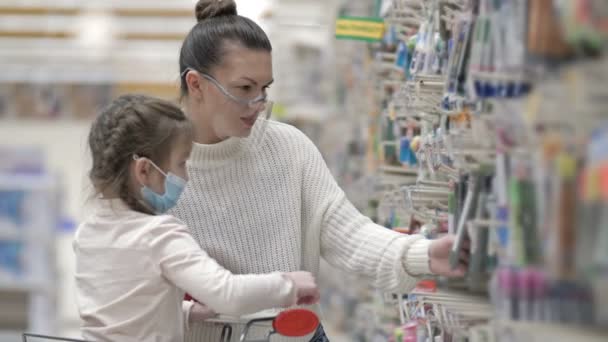 Moeder en kind met beschermende maskers kiezen briefpapier in de supermarkt. Winkelen met kinderen tijdens de virusuitbraak. — Stockvideo