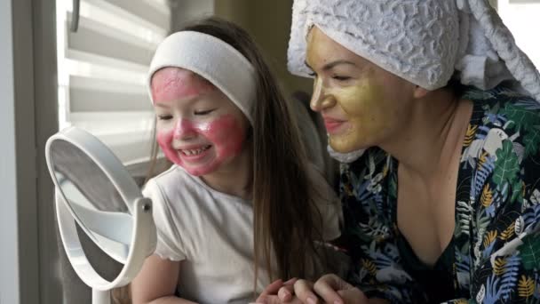 Giovane donna con sua figlia 6-7 anni ha applicato maschere cosmetiche ai loro volti. Si guardano allo specchio e ridono allegramente. — Video Stock