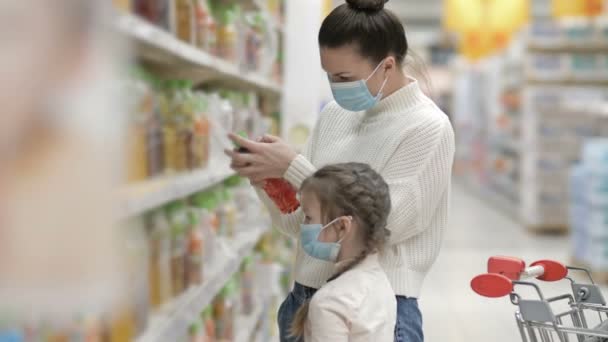 Anya és lánya, 6-7 éves, válasszanak gyümölcslevet a szupermarketben. Mindkettő védőmaszkot visel. Vásárlás gyermekekkel a vírus kitörése alatt. — Stock videók