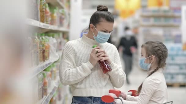 Mamma och dotter, 6-7 år, väljer juice i snabbköpet. Båda bär skyddsmasker. Shopping med barn under virusutbrottet. — Stockvideo