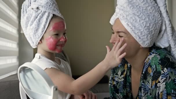 La niña con una máscara cosmética en la cara ayuda a su madre a hacer una máscara cosmética. Felices juntos. — Vídeos de Stock