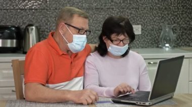 Koruyucu maskeli bir çift online alışveriş için evde dizüstü bilgisayar kullanıyor. Koronavirüs salgını sırasında alışveriş..