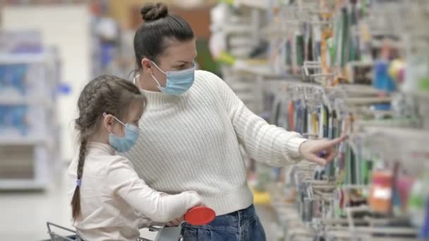 Koruyucu maskeler takan anne ve çocuk süpermarketten kırtasiye malzemesi seçiyorlar. Virüs salgını sırasında çocuklarla alışveriş yapmak. — Stok video