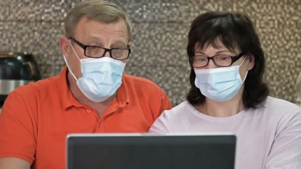 Pareja senior en máscaras protectoras usando portátil en casa para compras en línea. Compras durante la pandemia del coronavirus. — Vídeo de stock