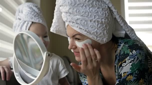 Schöne Mutter im Bademantel und ihre kleine Tochter zusammen mit Handtüchern auf dem Kopf und Maske auf dem Gesicht, während sie Wellness-Behandlungen genießt. — Stockvideo