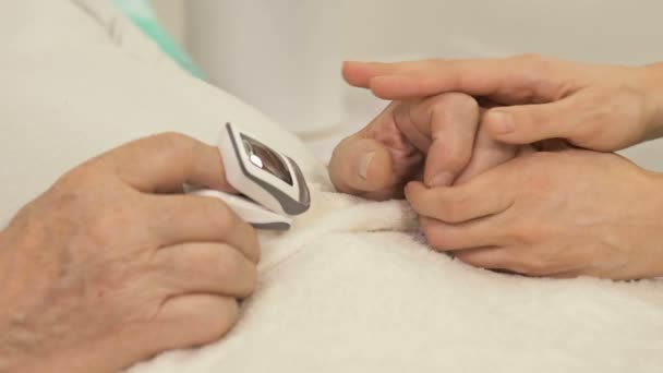 La mano femenina sostiene la mano de un paciente anciano con síntomas graves acostado en una cama en un hospital de emergencia. gesto de apoyo. — Vídeos de Stock