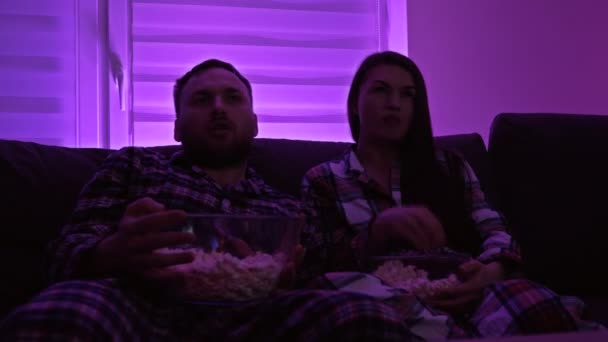 Casamento casal está sentado no sofá de pijama e assistindo com interesse o que está acontecendo na tela da TV. Eles comem pipocas ao mesmo tempo.. — Vídeo de Stock