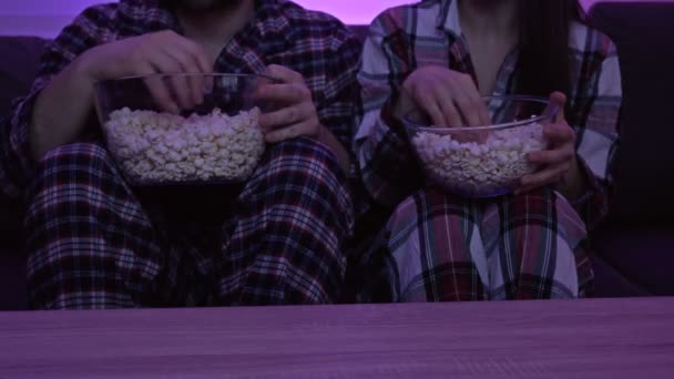 Pareja casada en pijama sentada frente al televisor y comiendo palomitas de maíz. — Vídeo de stock