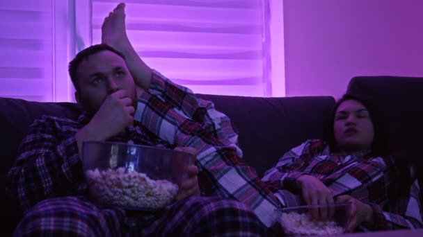 La pareja casada está viendo televisión y comiendo palomitas de maíz. Cuarentena domiciliaria durante la pandemia de Covid-19. — Vídeos de Stock