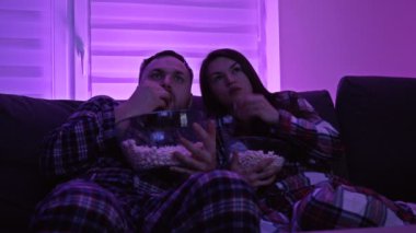 Kanepede yan yana oturmuş patlamış mısır yiyen ve geceleri korku filmi izleyen güzel bir çift. Covid-19 salgını sırasında ev karantinası.