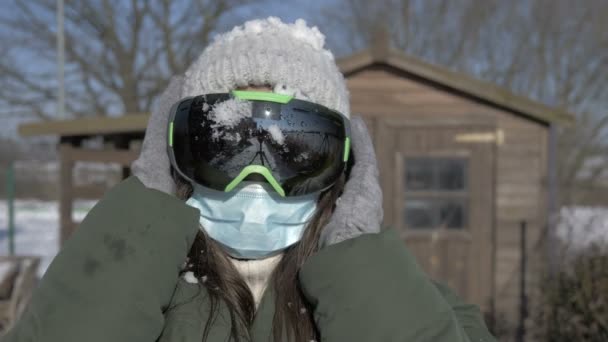 Güneşli bir kış gününde COVID-19 Coronavirus sırasında kayak gözlüğü ve tıbbi maske takan genç bir kadının portresi. Kadın gözlüklerini ve maskesini çıkarıyor.. — Stok video