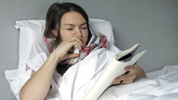 Zieke jonge vrouw ligt in bed met griep of coronavirus symptomen. De vrouw is aan de telefoon aan het praten, schrijft iets en spuit medicijnen in haar neus. Kenmerken van werken op afstand. — Stockvideo