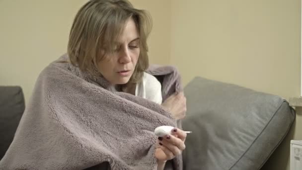 Młoda kobieta siedzi owinięta w koc i mierzy temperaturę. Ma objawy grypy, przeziębienia lub koronawirusu.. — Wideo stockowe