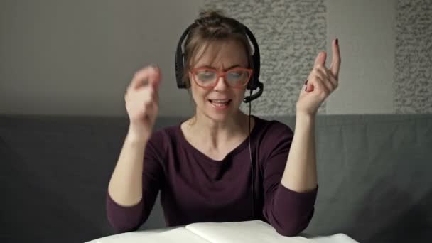 Radosna kobieta siedzi przy stole roboczym. Śpiewa i tańczy do rytmu muzyki grającej przez słuchawki. — Wideo stockowe