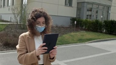 Koruyucu maskeli, elinde tabletle ıssız bir caddede yürüyen güzel bir kadın. Covid-19 salgınından dolayı tecrit.