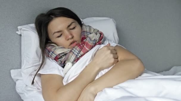 Mujer joven enferma se acuesta en la cama y mide la temperatura. Tiene tos, escalofríos, secreción nasal. Síntomas de gripe, resfriado o coronavirus. — Vídeos de Stock