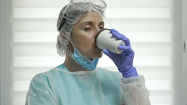Koruyucu kıyafetli yorgun kadın doktor ya da hemşire kahve içiyor. Salgın Covid-19.