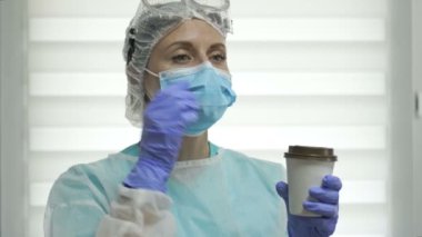 Koruyucu kıyafetli yorgun kadın doktor ya da hemşire kahve içiyor. Salgın Covid-19.