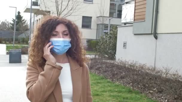 Bella donna in maschera protettiva cammina lungo una strada deserta della città e parla su un telefono cellulare. Blocco a causa della pandemia di Covid-19. — Video Stock