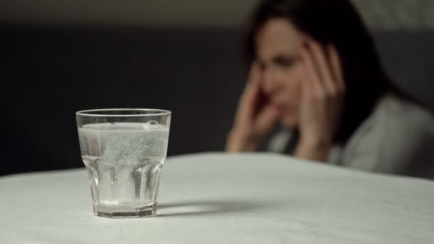 Ett glas vatten med en upplöst tablett. Närbild. I bakgrunden gnuggar en plågad kvinna sina tempel.. — Stockvideo
