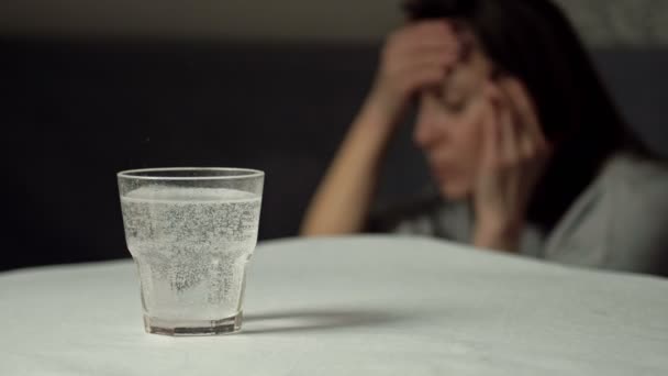 Un vaso de agua con una tableta disolvente. Primer plano. Al fondo, una mujer dolorida se frota las sienes. — Vídeo de stock